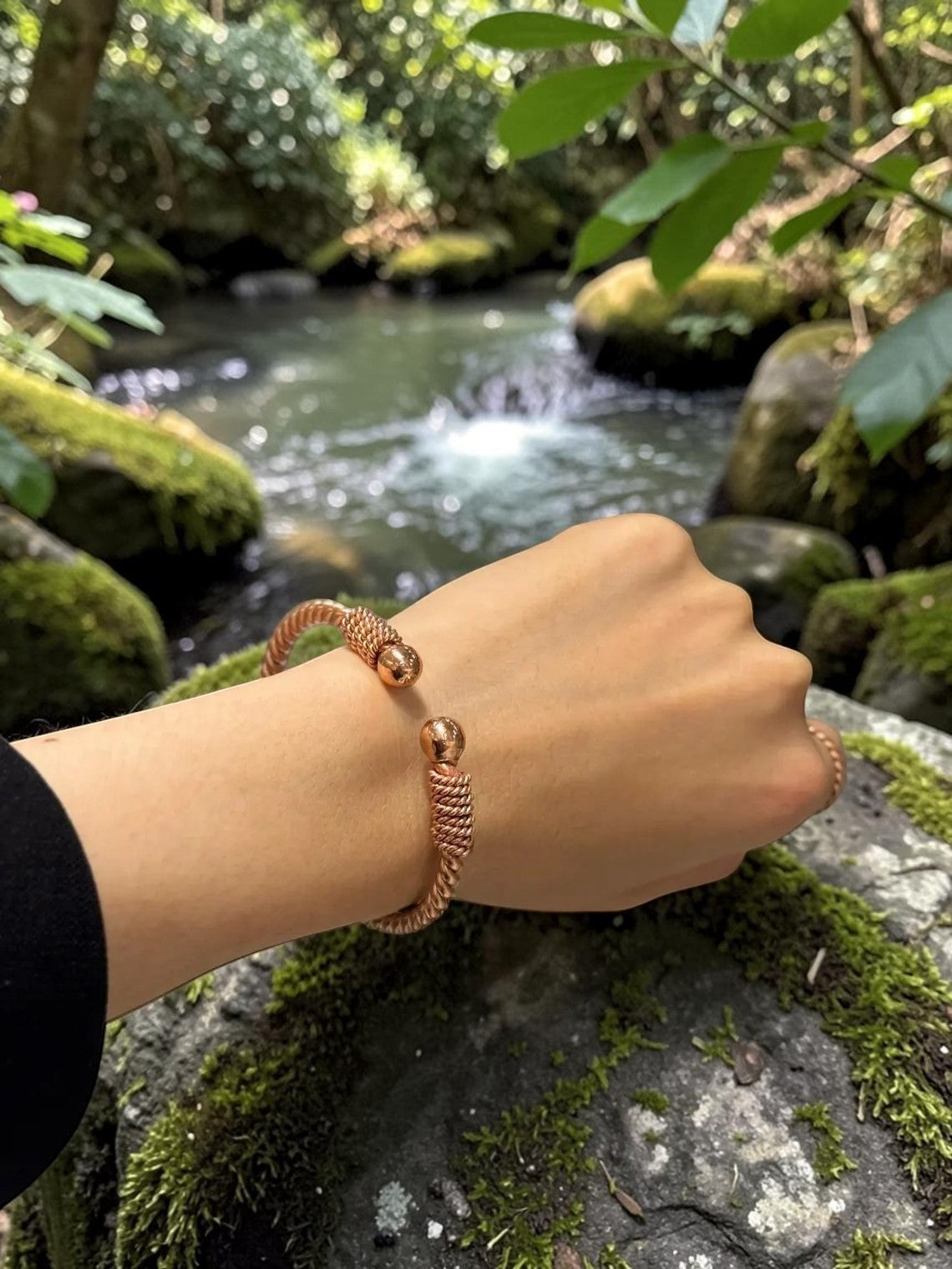 Vòng đeo tay bản hẹp bằng đồng nguyên chất màu vàng hồng, thiết kế xoắn với hai quả cầu nhỏ ở hai đầu, đeo trên cổ tay người mẫu trong khung cảnh thiên nhiên.