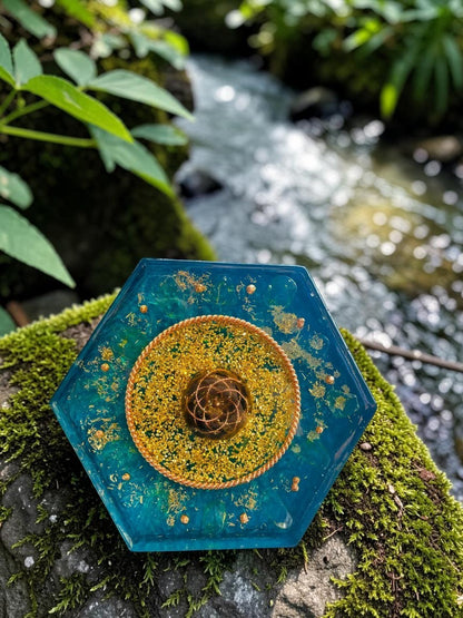 Tháp orgonite hình lục giác màu xanh ngọc bích với các hạt vàng lấp lánh và trung tâm hình xoắn ốc bằng đồng nguyên chất, được đặt trên đá phủ rêu bên cạnh dòng suối.
