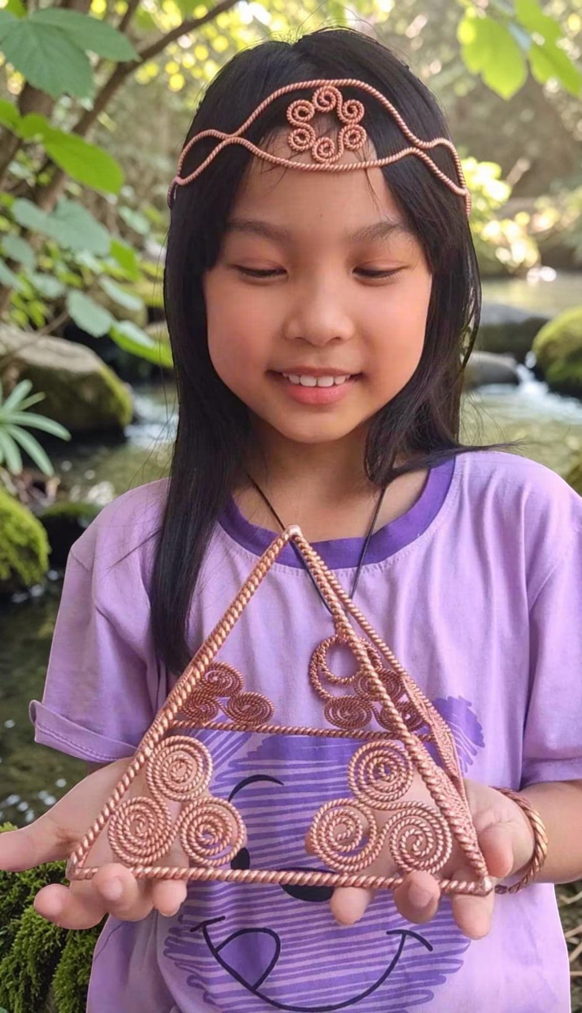 Cô bé châu Á đội vương miện đồng xoắn và cầm một đĩa orgone hình tam giác bằng đồng nguyên chất 99,9% với tần số 177MHz.