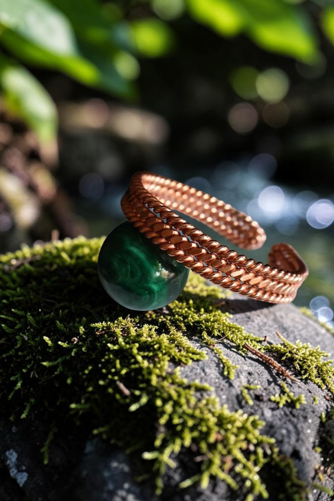 Vòng tay đồng nguyên chất abusta.viking với thiết kế xoắn ốc, đặt trên quả cầu đá malachite xanh lá cây, trên nền đá phủ rêu và dòng nước chảy.
