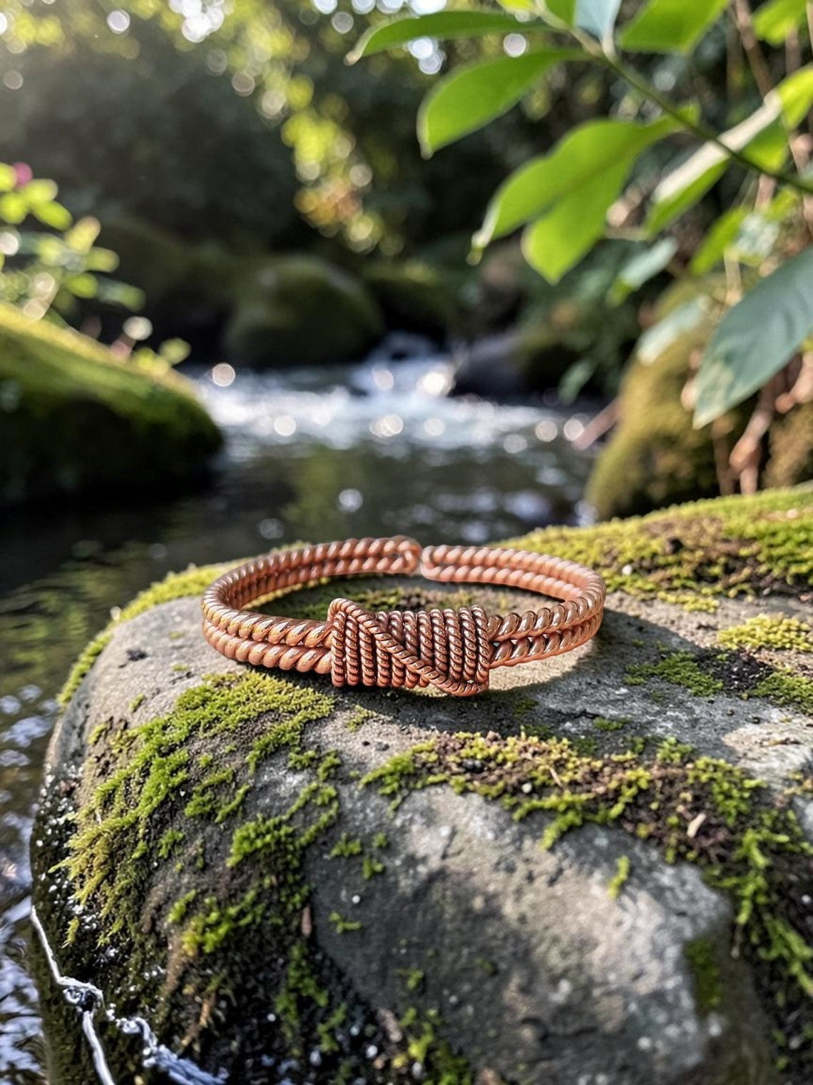 Vòng tay đồng đỏ thủ công abusta.viking với thiết kế xoắn và nút thắt tinh xảo, đặt trên tảng đá phủ rêu bên bờ suối.