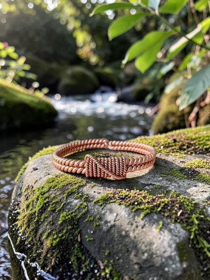 Vòng tay đồng đỏ thủ công abusta.viking với thiết kế xoắn và nút thắt tinh xảo, đặt trên tảng đá phủ rêu bên bờ suối.