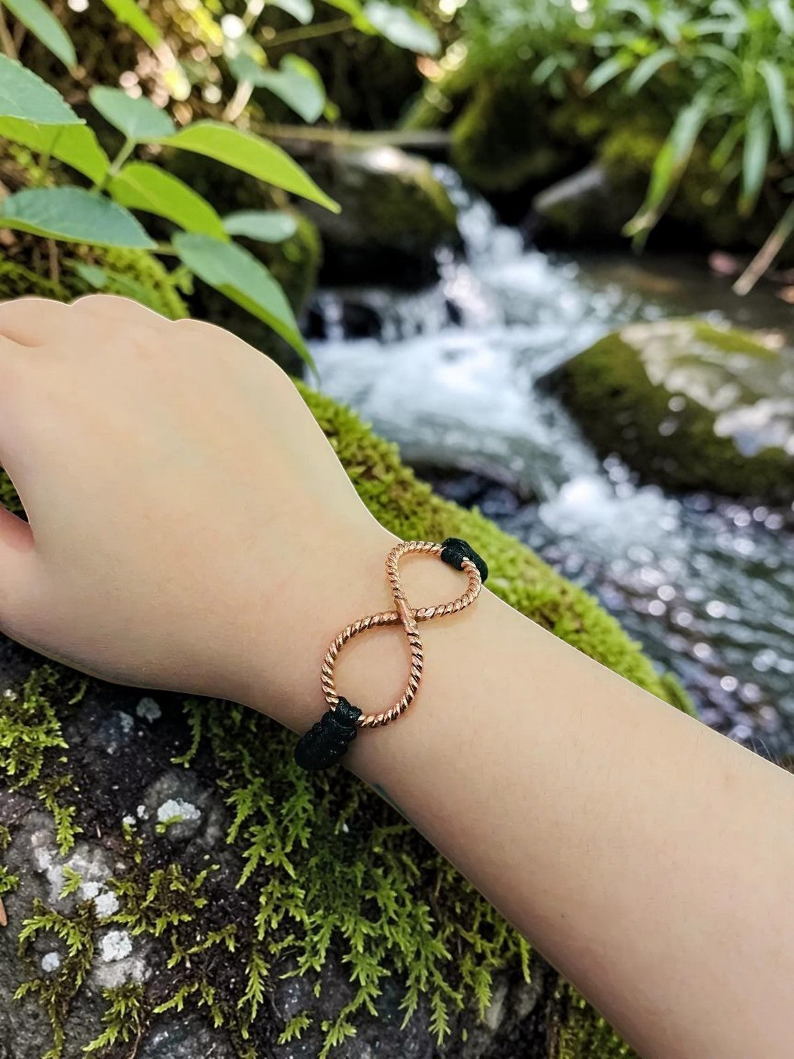 Vòng đeo tay Torus Orgonite Rodin Coil bằng đồng nguyên chất, thiết kế hình số 8 xoắn, điểm nhấn màu đen, trên tay người mẫu với bối cảnh suối nước chảy tự nhiên.