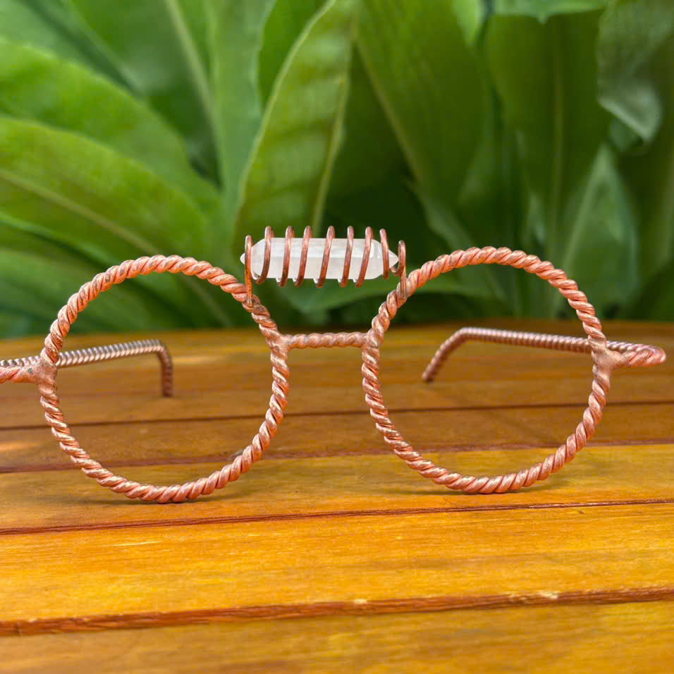 Handmade clear quartz copper tensor glasses with twisted round wire-frame rims, on a wooden surface with green leaves background