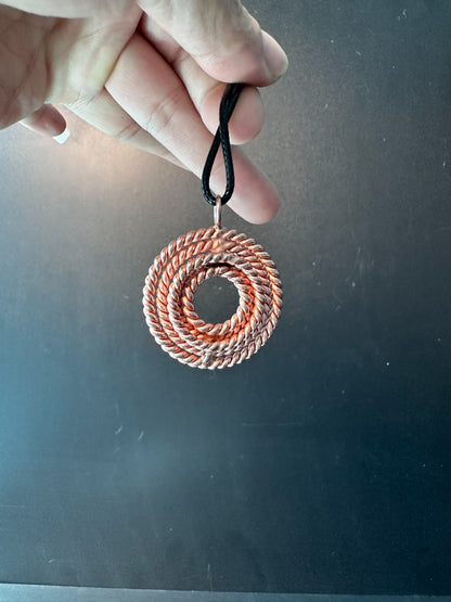 Copper five-frequency tensor pendant held by its black cord, showing the hand-woven concentric coil