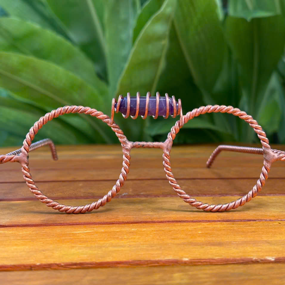 Handmade twisted copper tensor glasses with round wire-frame lenses and a striped stone nose bridge, on a wooden table with green tropical leaves background