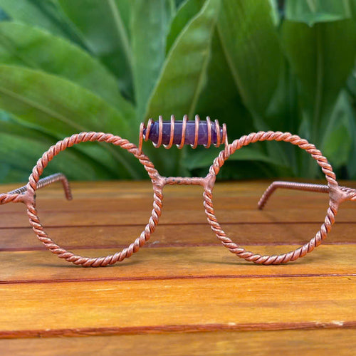 Handmade twisted copper tensor glasses with round wire-frame lenses and a striped stone nose bridge, on a wooden table with green tropical leaves background