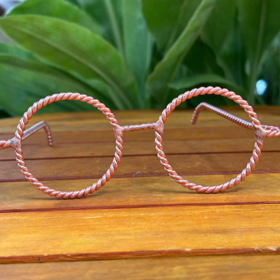 Handmade twisted copper tensor glasses with round wire-frame lenses, resting on a wooden table with blurred green plant background