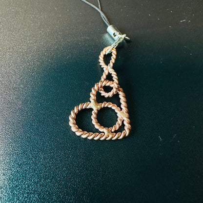 Handmade copper phone charm shaped like a heart from twisted tensor wire, on a strap against a dark surface