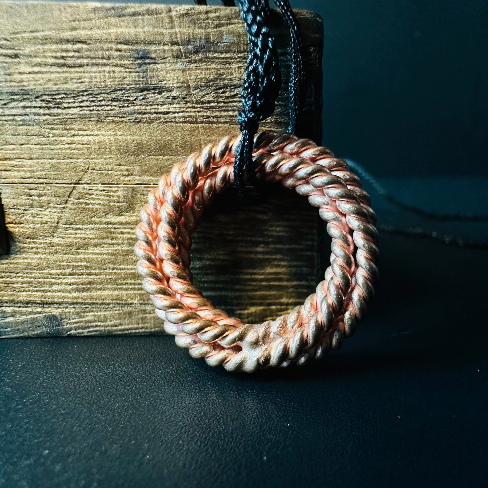 Handmade copper tensor ring pendant, double-twisted closed circle on a black cord, leaning against rustic wood