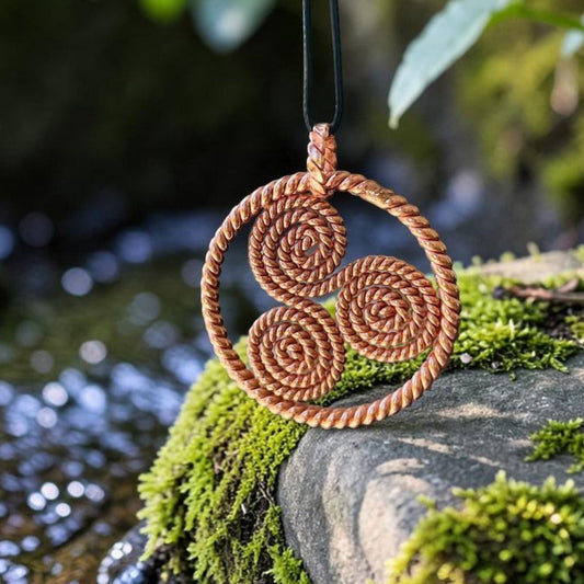 Copper triskelion pendant with three spirals on a black cord, resting on a mossy rock by a stream