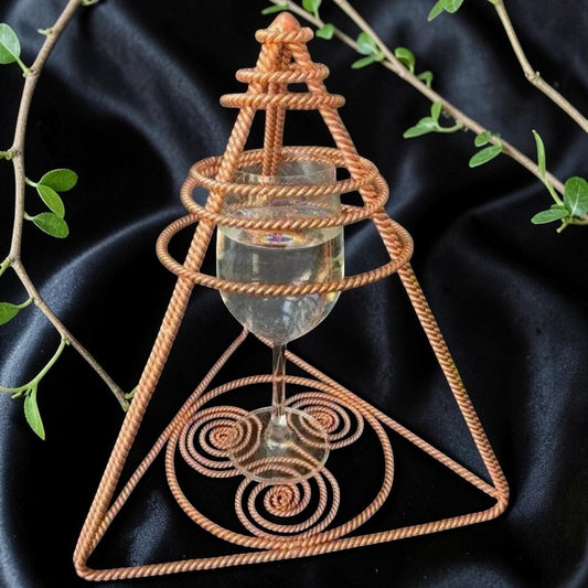 Handmade copper water charging pyramid with a central glass and twin spiral coils, on dark fabric
