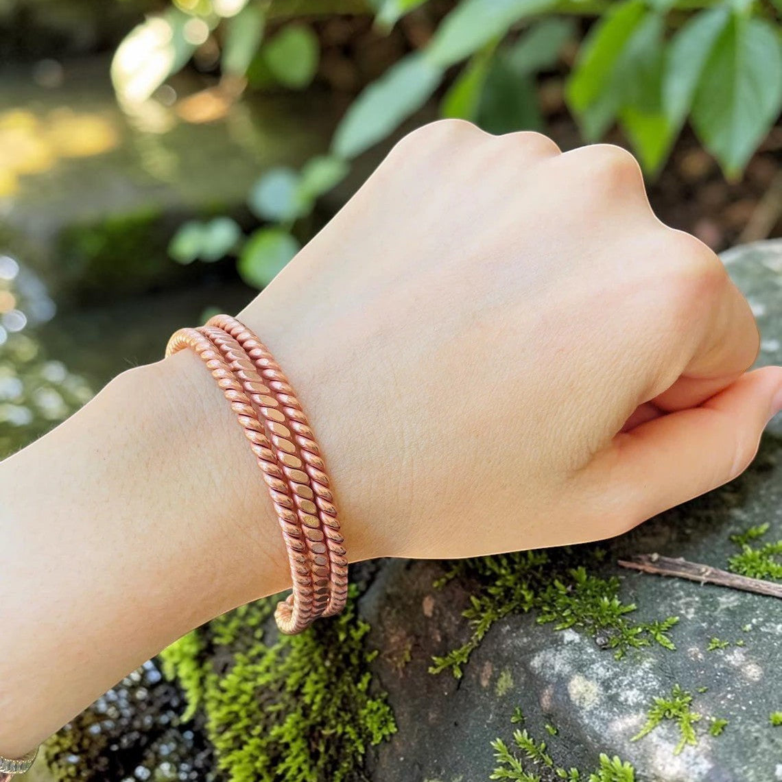 Two double twist copper tensor bracelets resting on a mossy rock by a stream, handmade open cuffs