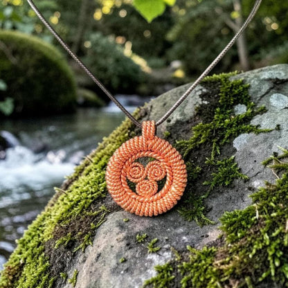 Nuban copper sacred geometry pendant with a triple spiral face, hanging on a cord over a mossy rock